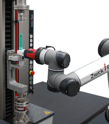 Robot de construcción ligera roboTest N posiciona una jeringa en la máquina de ensayos de materiales y lleva a cabo el ensayo automatizado